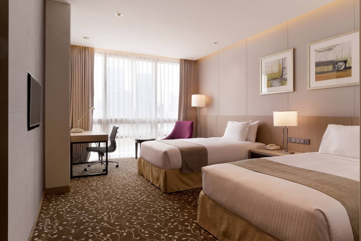 A hotel room with two beds, a desk, a chair, a lamp, and a window with curtains.