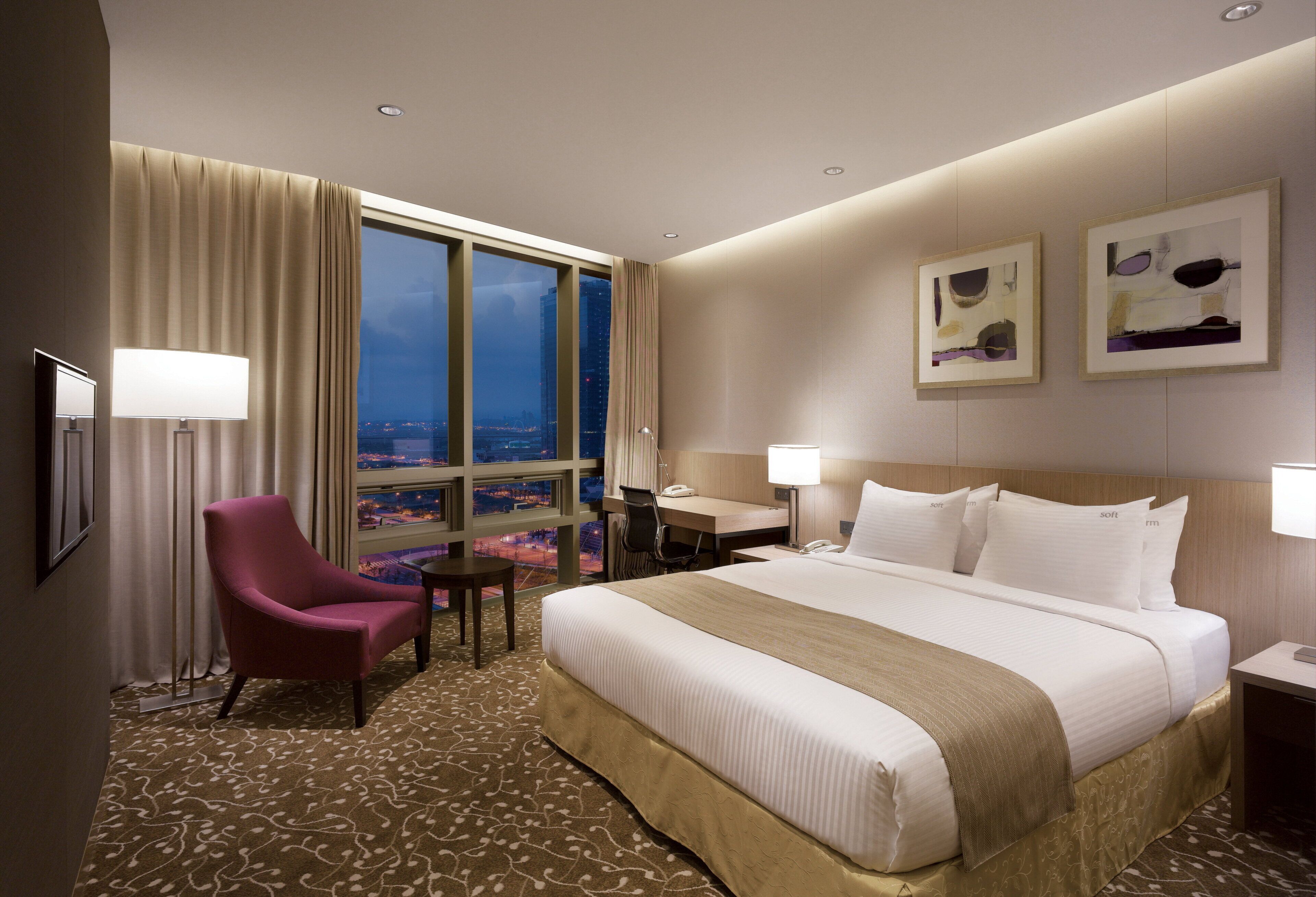 Ein Hotelzimmer mit einem großen Bett, einem Schreibtisch, einem Stuhl und Blick auf die beleuchtete Stadt.