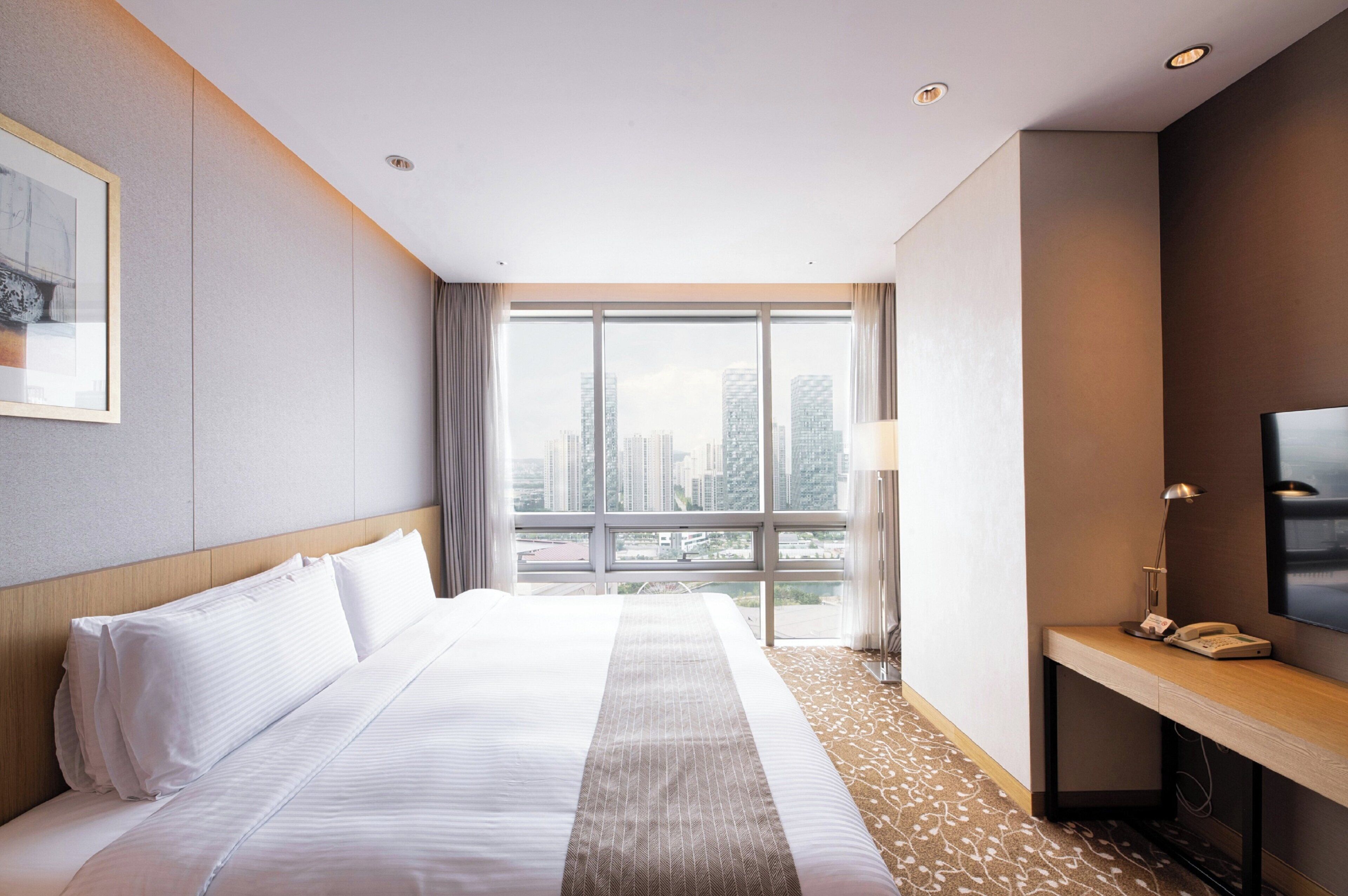 A hotel room with a large bed, a desk with a lamp, a TV, and a city view.
