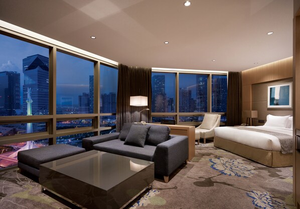 A modern hotel room with a large city view, a sofa, a bed, and a desk.