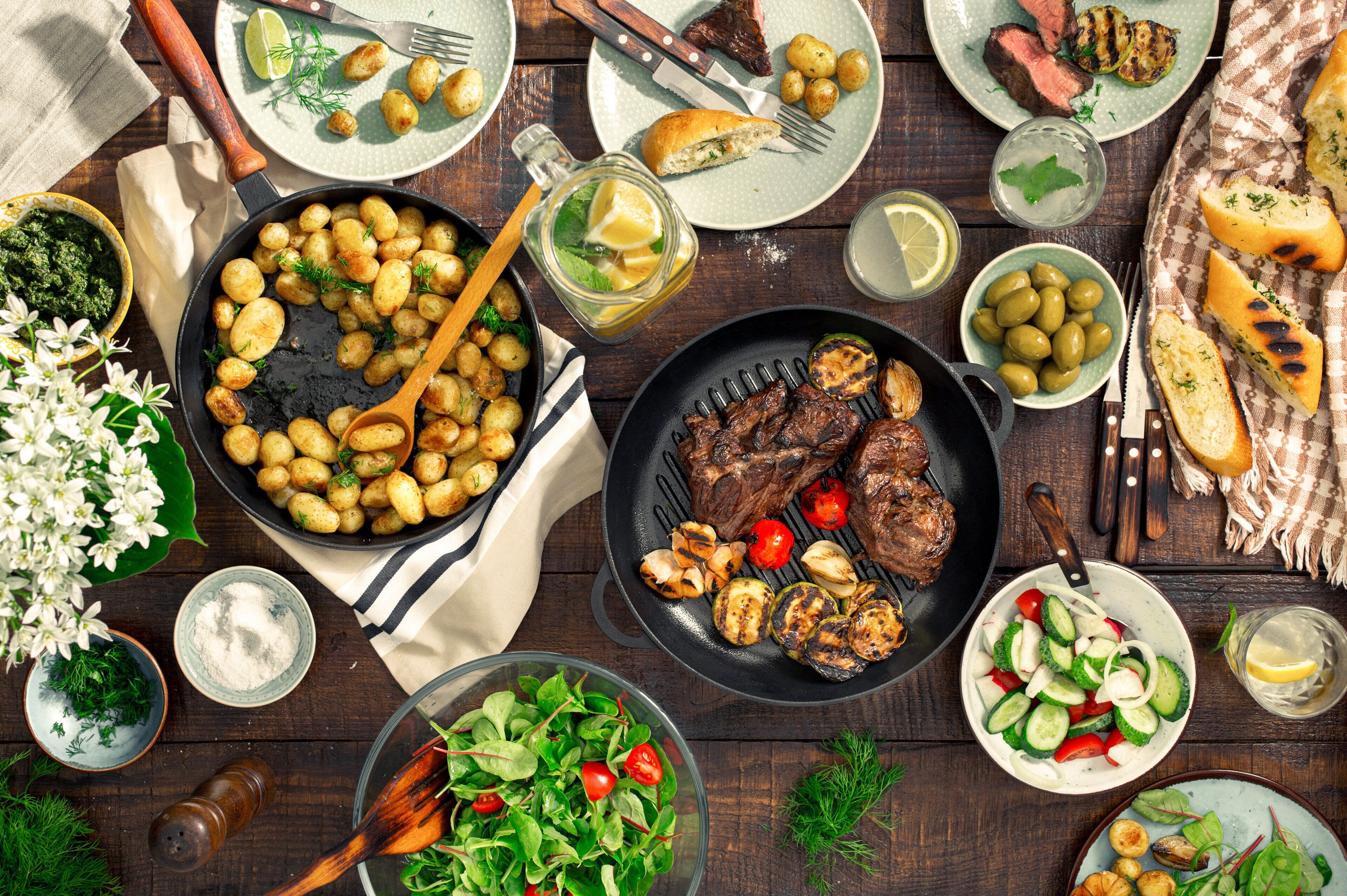 Diversos pratos, incluindo carne grelhada, batatas assadas e uma salada, indicando um ambiente de refeição.