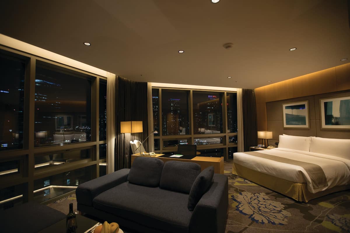 A hotel room with a large window, a bed, a sofa, a desk with a laptop, and a nightstand with a lamp.