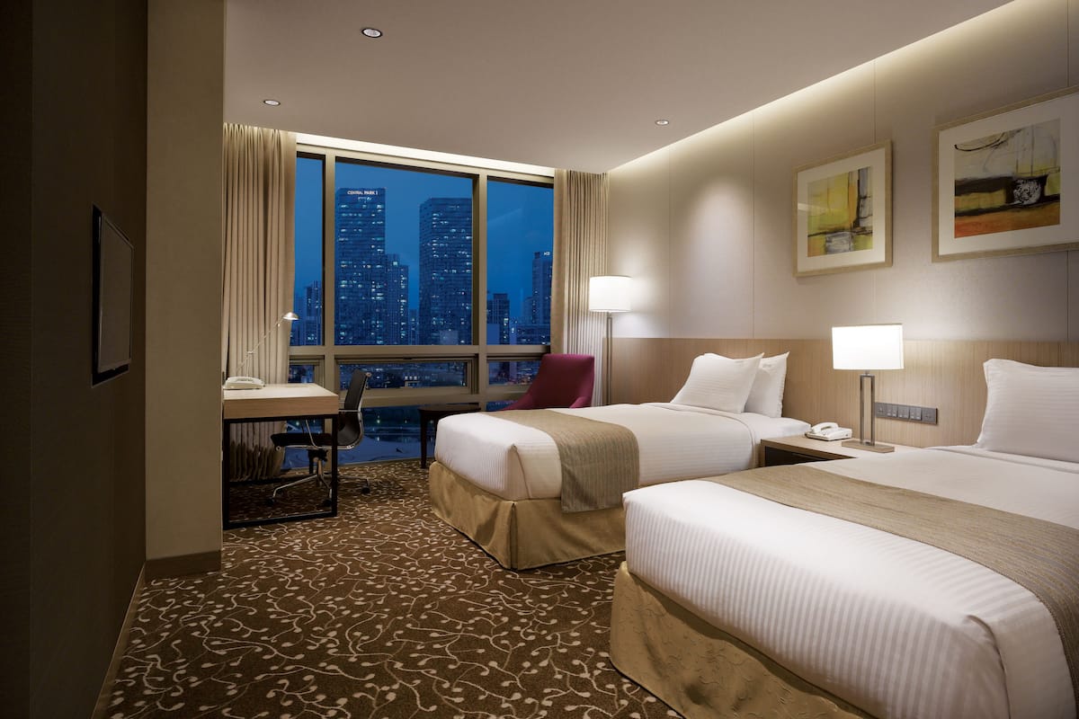 A hotel room with two beds, a desk, a chair, and a large window with a city view.