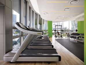 A gym with treadmills, weights, and a view of a cityscape through large windows.