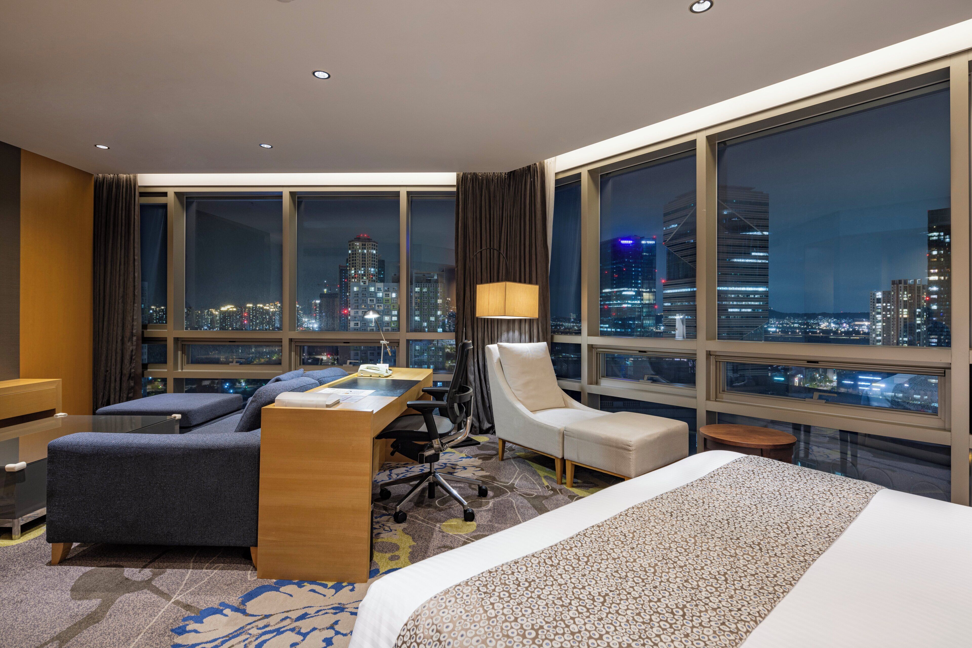 A hotel room with a large bed, a desk, a chair, and a sofa. There is a view of a cityscape through large windows.