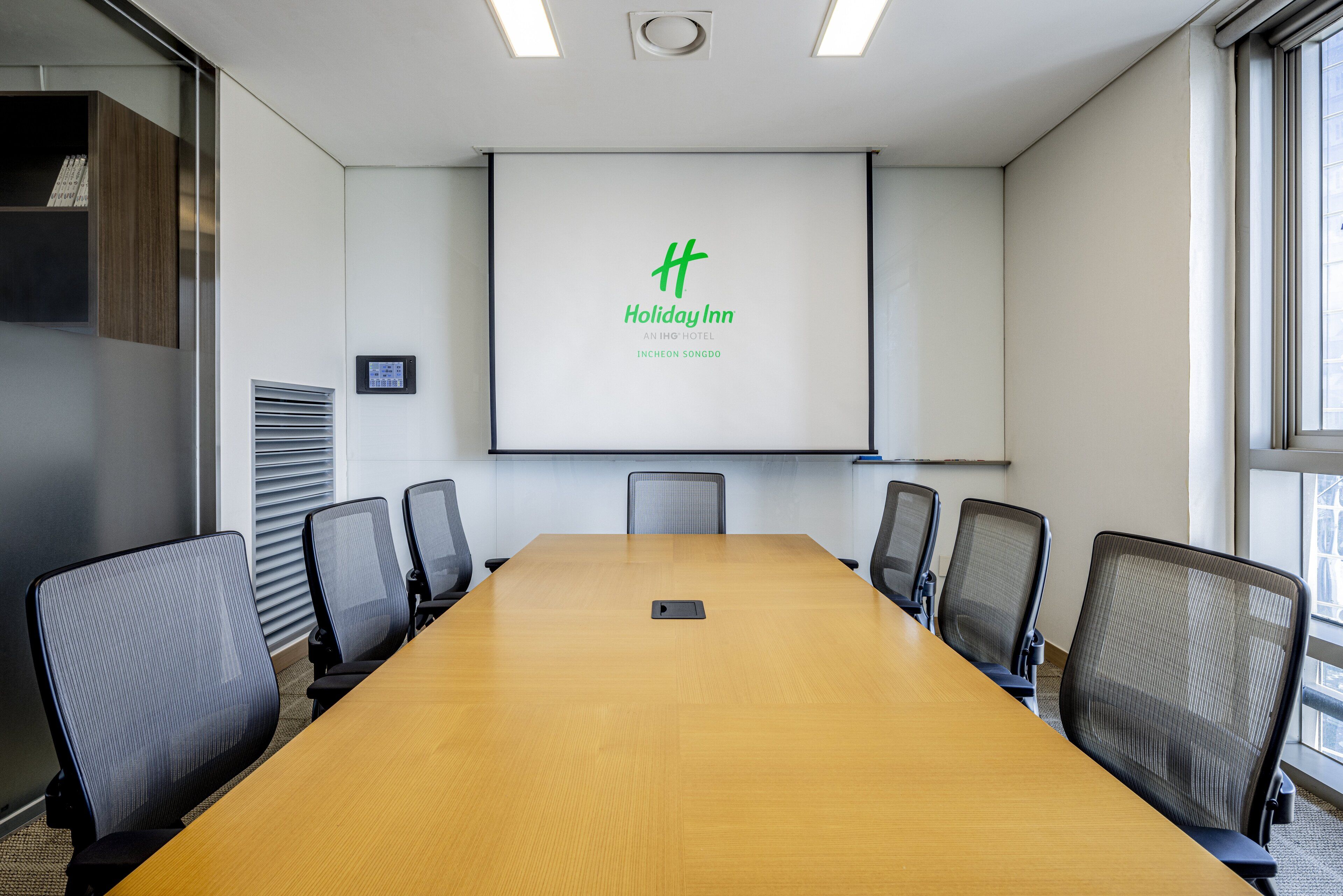 A conference room with a long table, six chairs, a projector screen displaying the Holiday Inn logo, and a window on the right.