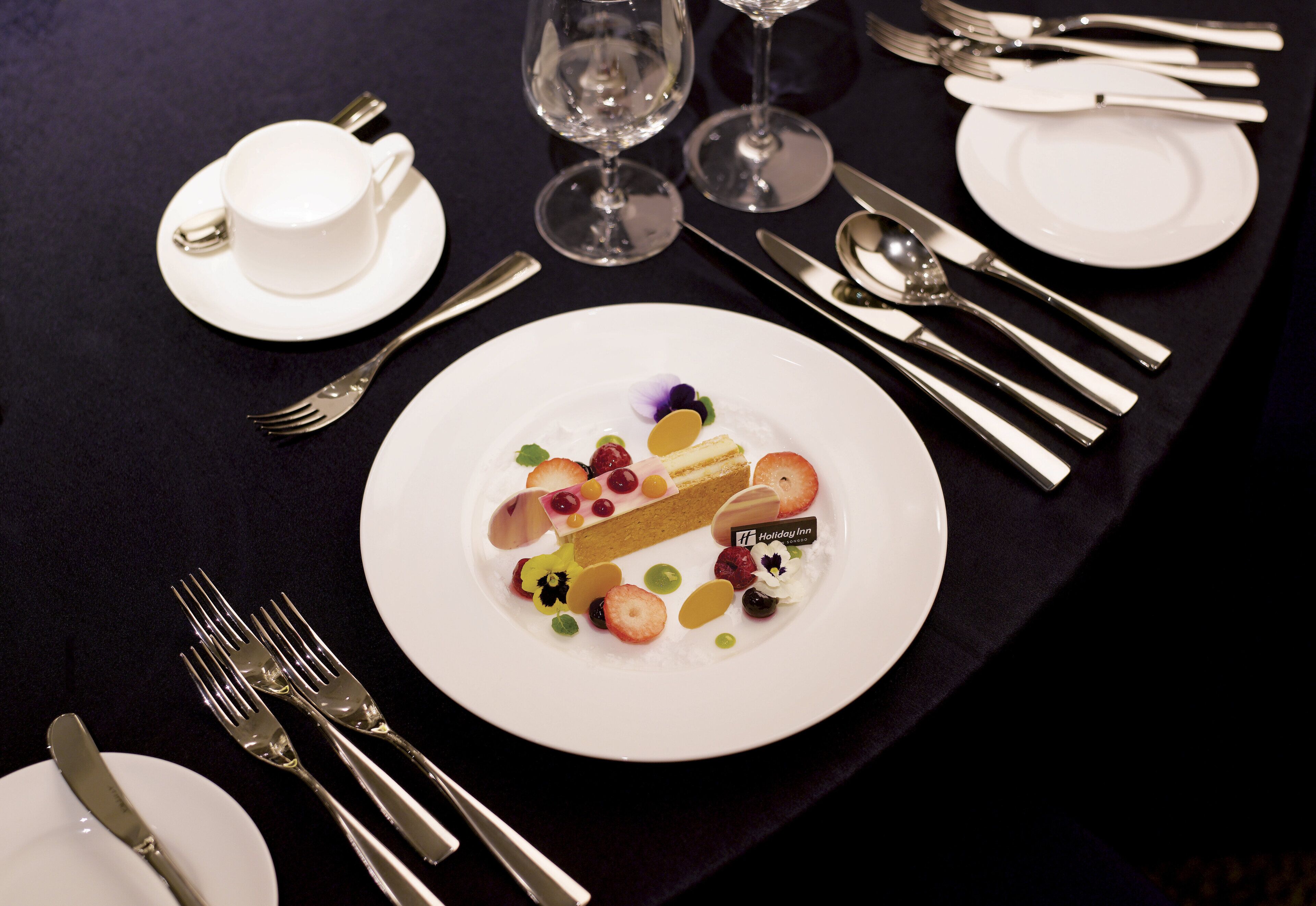 A table set with white plates, silverware, and a dessert with fruit and a chocolate piece.