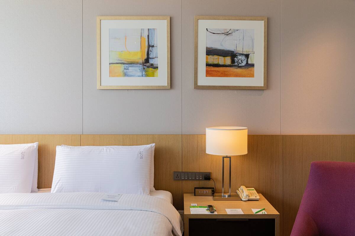 A hotel room with a bed, two framed paintings on the wall, a bedside lamp, a nightstand with a phone, and a purple chair.
