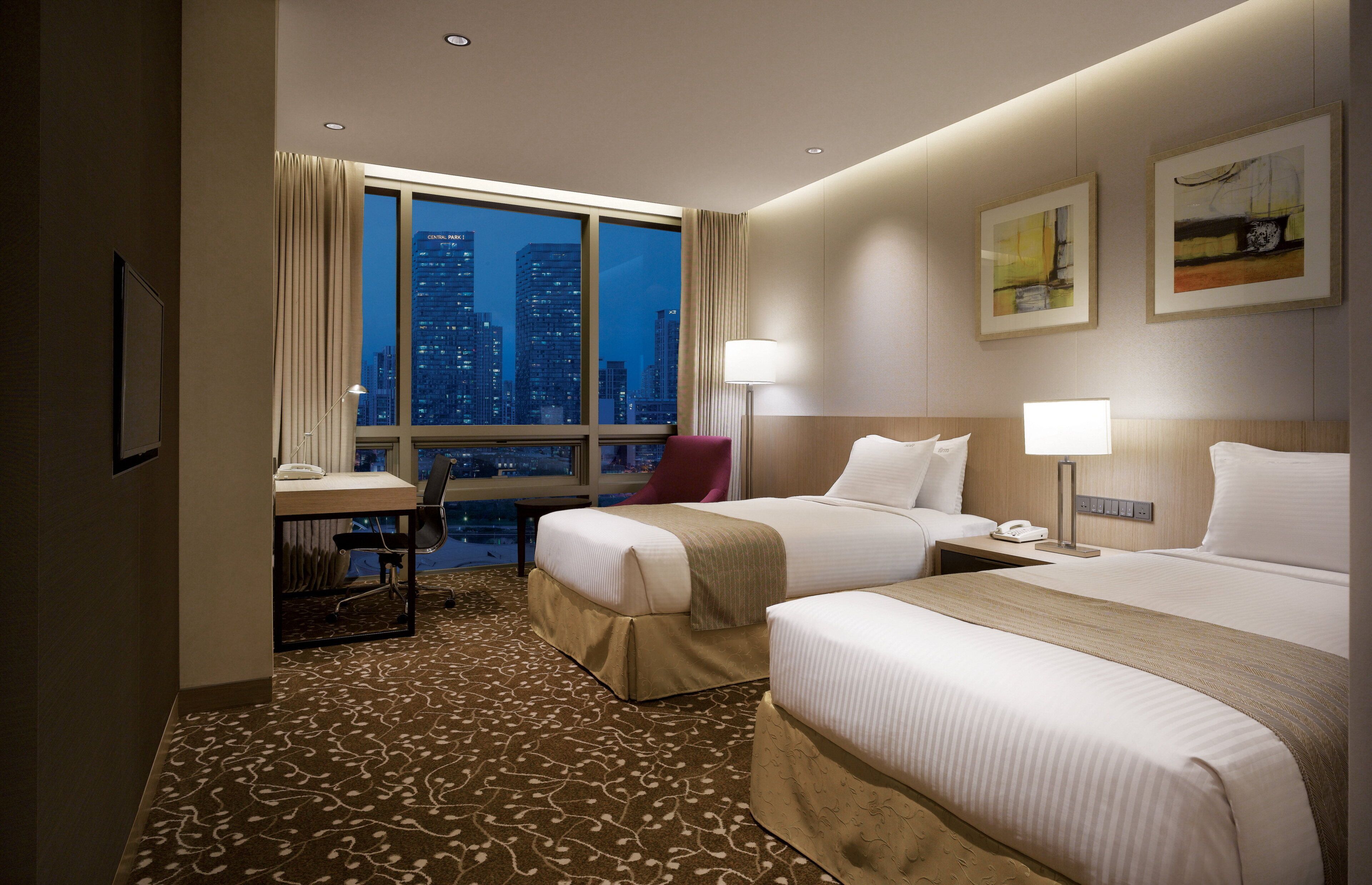A hotel room with two beds, a desk, a chair, a nightstand, and a view of the cityscape.