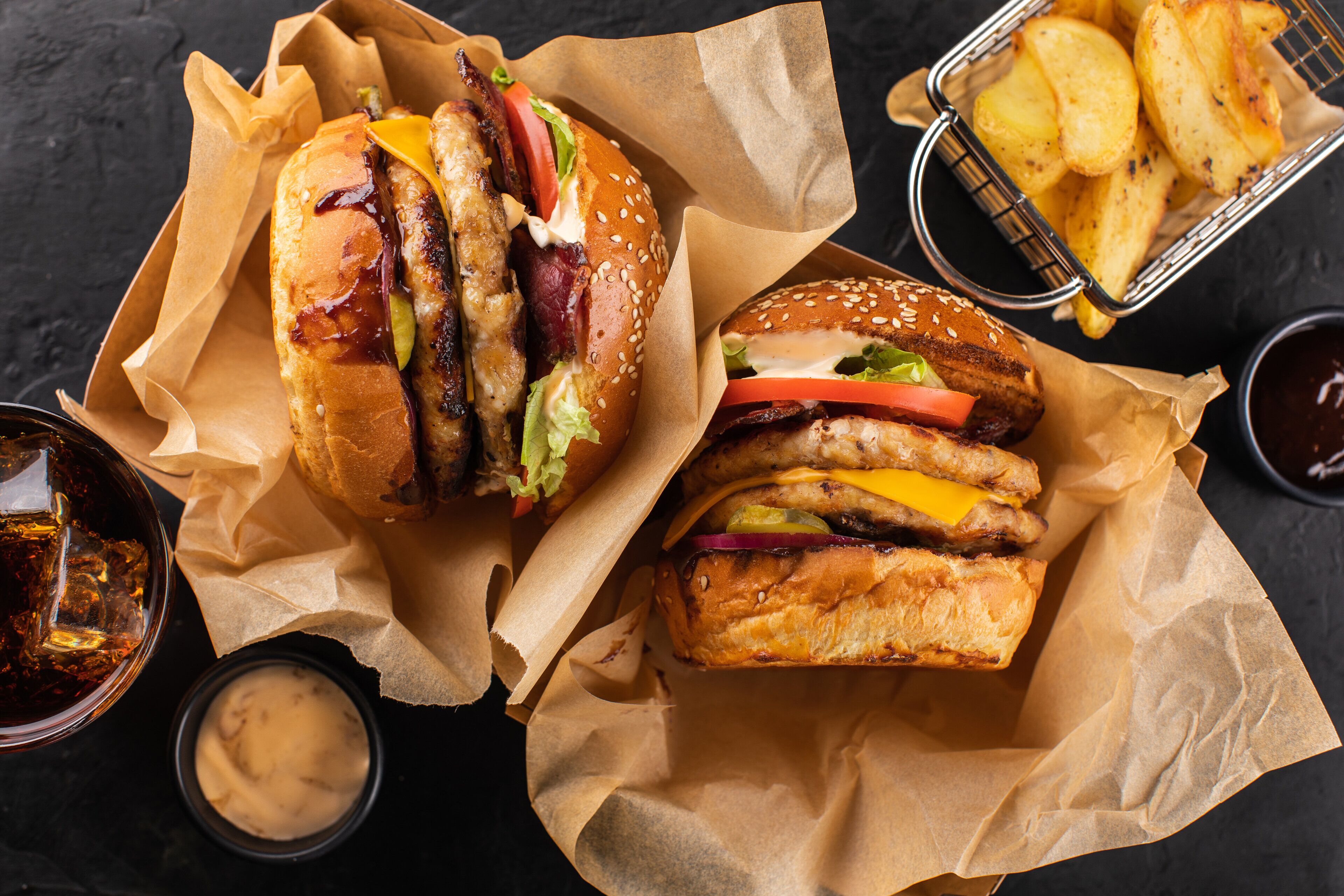 Two burgers with lettuce, tomato, and cheese, and a basket of fries on a dark surface.