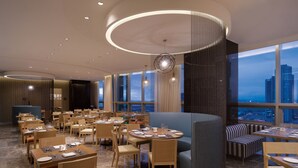A modern restaurant with a panoramic city view, featuring round tables, wooden chairs, and a stylish ceiling design. - Holiday Inn Incheon Songdo by IHG (Incheon)