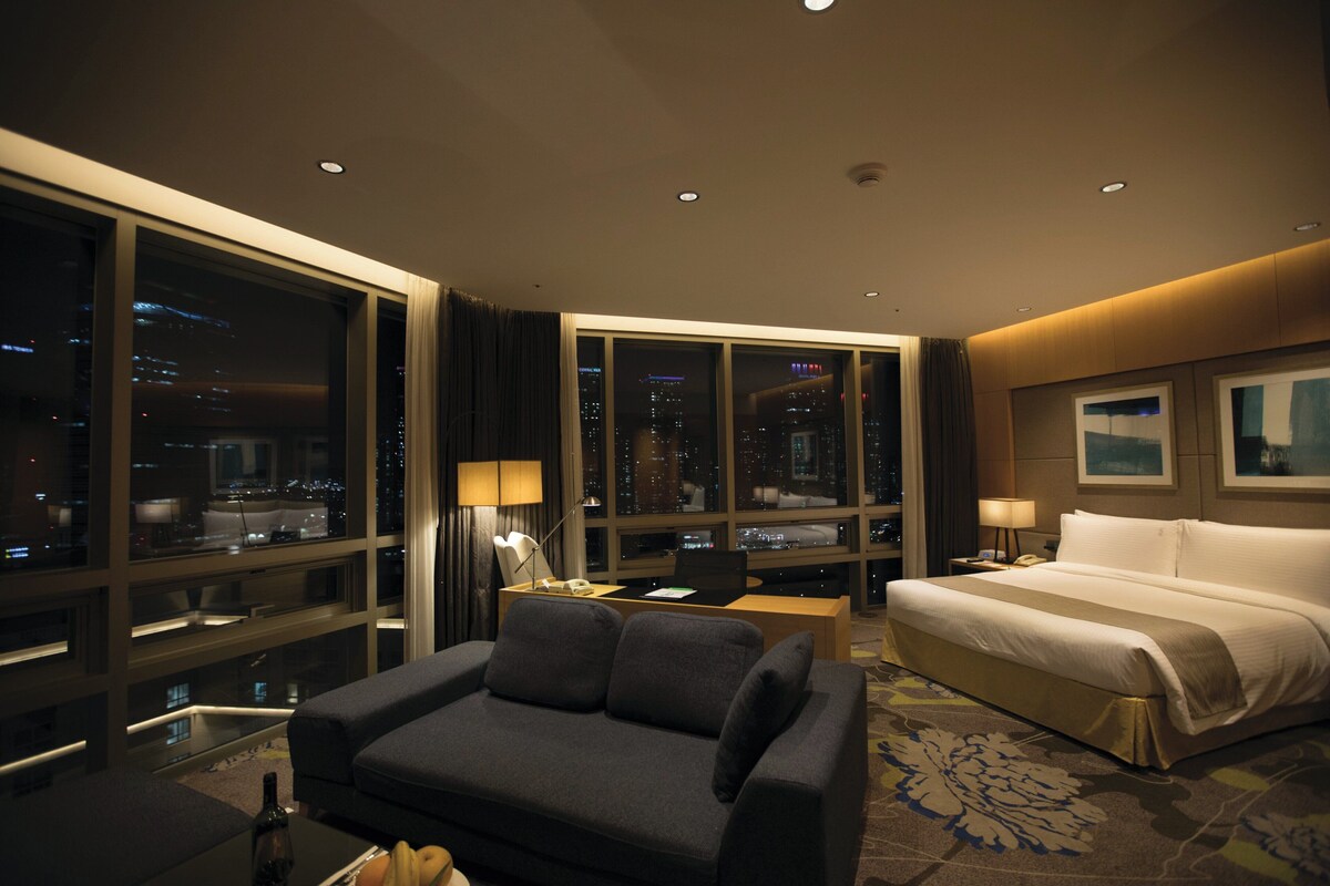 A hotel room with a large window, a bed, a sofa, a desk with a laptop, and a nightstand with a lamp.