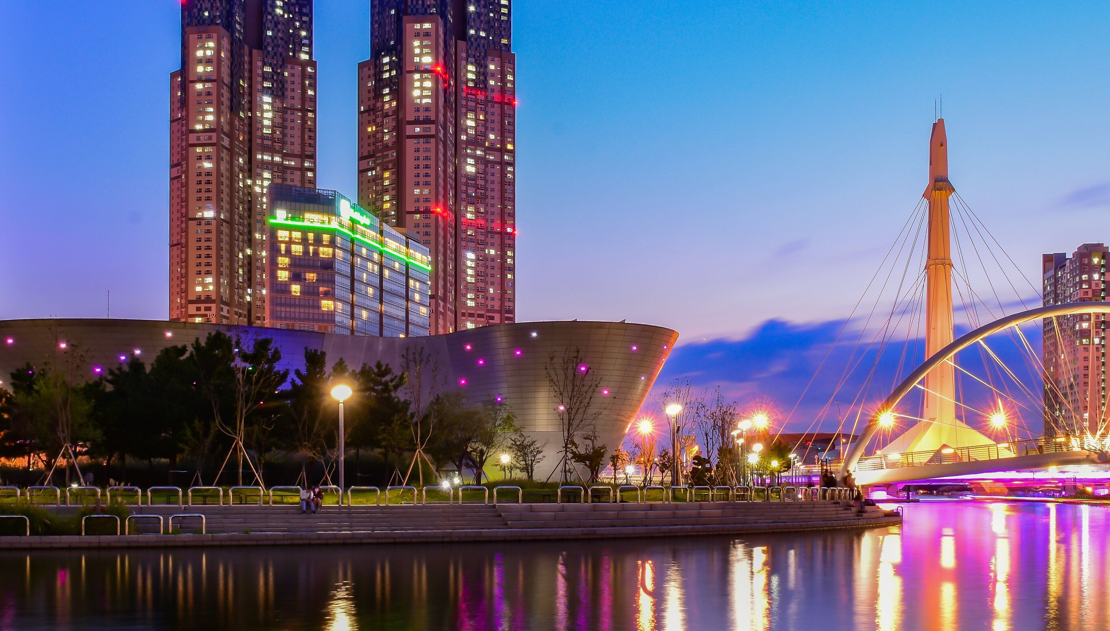 A cityscape with illuminated high-rise buildings, a bridge with a cable design, and a body of water reflecting the lights.