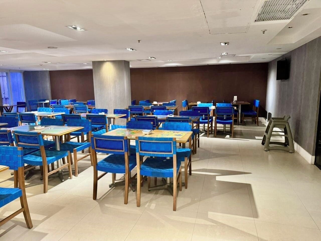 A spacious room with multiple tables and chairs arranged in rows, a large pillar, and a television mounted on the wall.