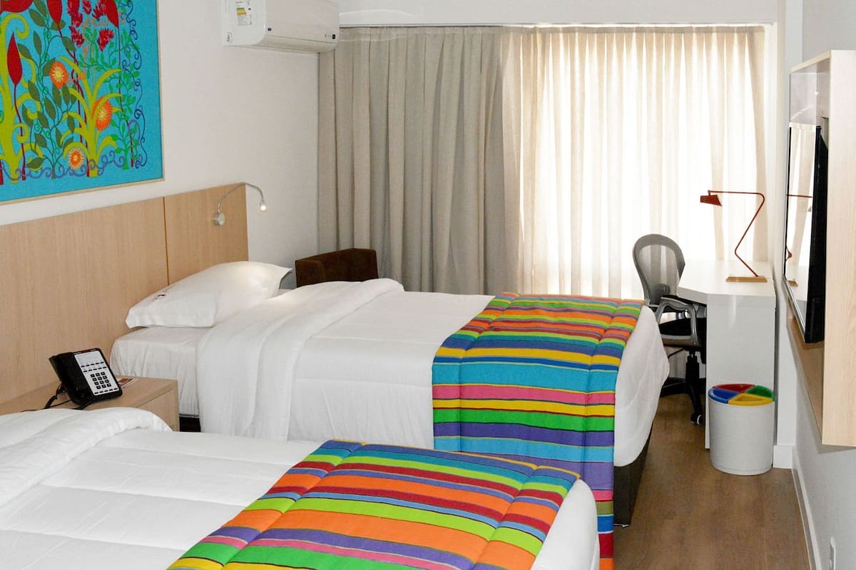 A hotel room with two beds, a desk, a chair, a TV, and a painting on the wall.