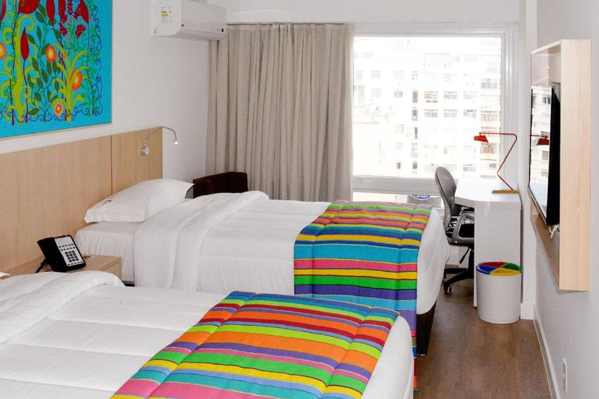 A hotel room with two beds, a desk, a chair, a TV, and a window with a view of buildings.