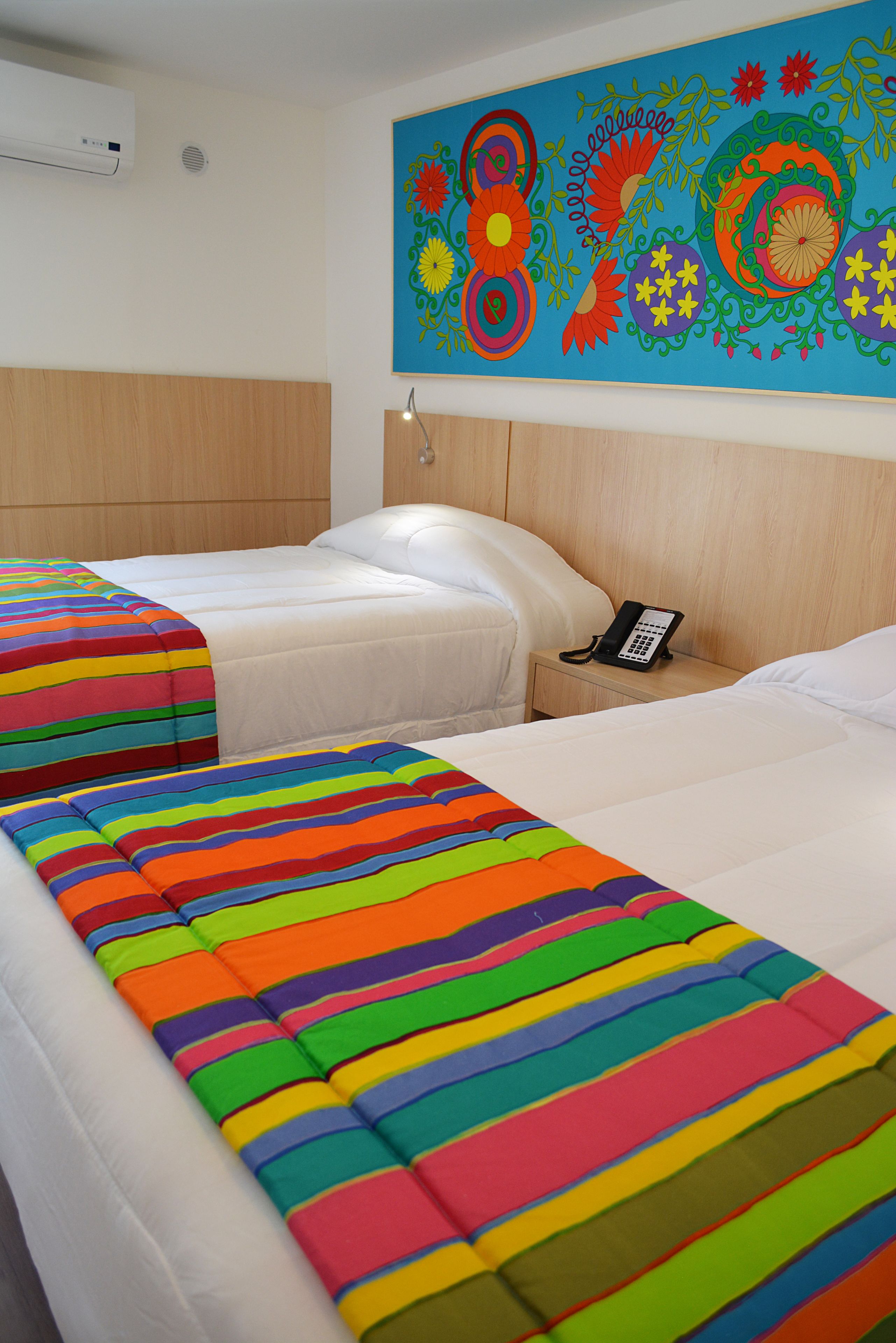 A hotel room with two beds, colorful striped blankets, a phone on the nightstand, and a vibrant floral wall mural.