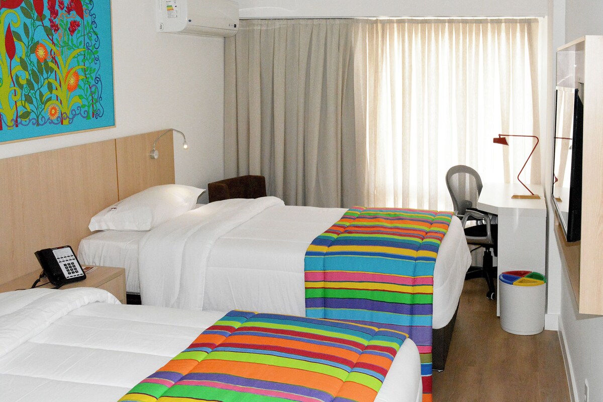 A hotel room with two beds, a desk, a chair, a television, and a telephone.