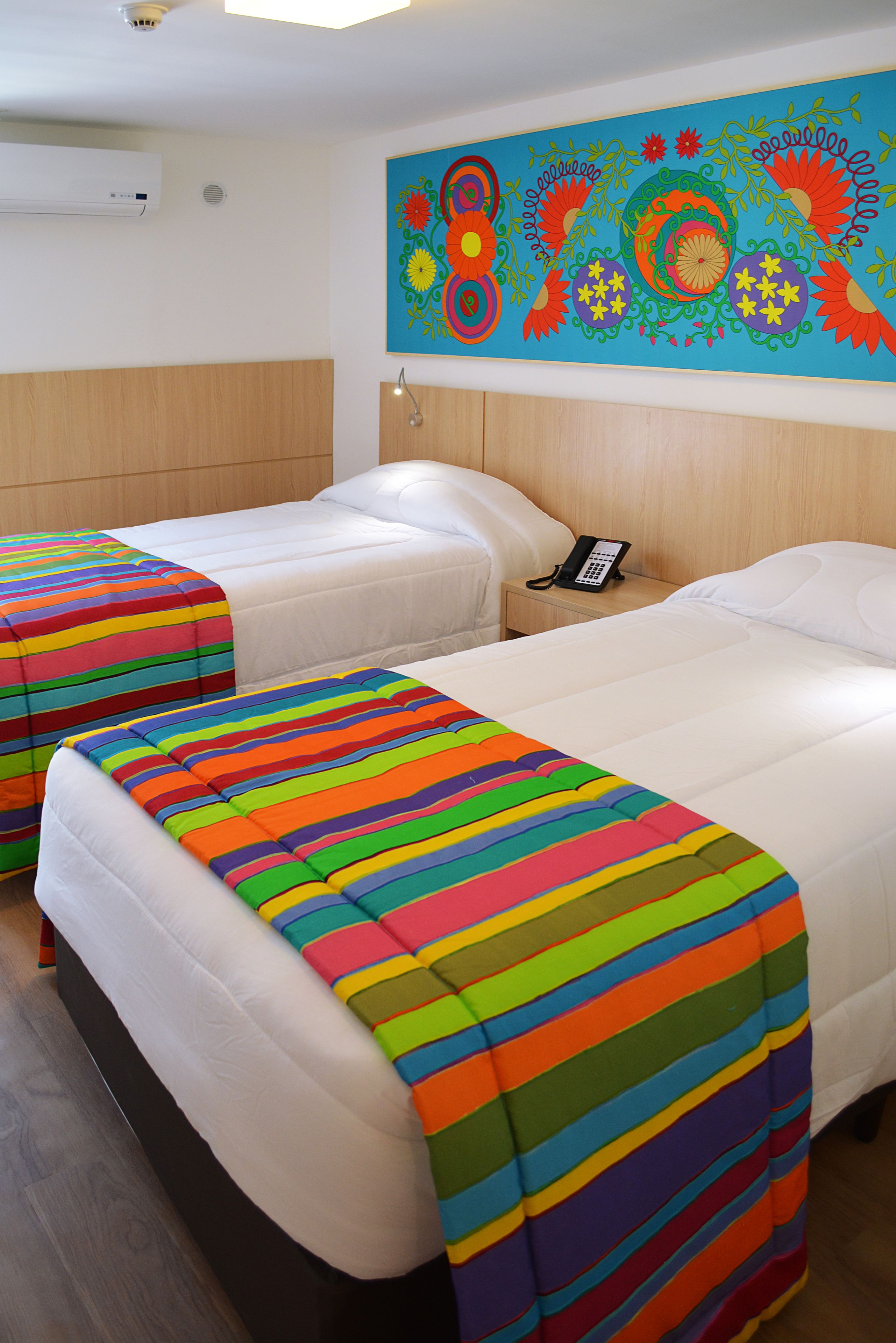 Two single beds with colorful striped bedspreads, a wooden headboard, a wall-mounted phone, and a decorative painting.
