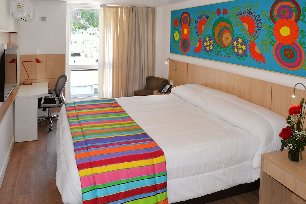 A hotel room with a large bed, a desk with a chair, a TV, and a colorful painting on the wall.