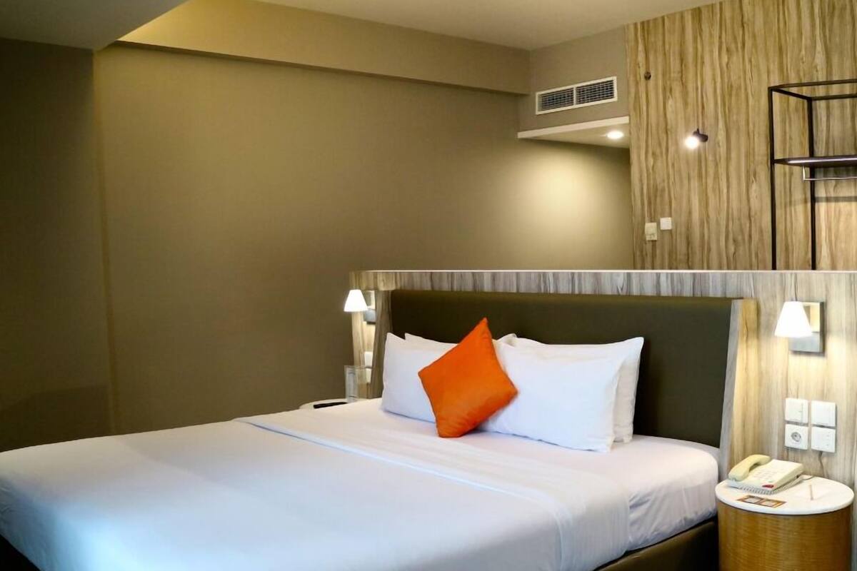 A hotel room with a bed, a bedside lamp, a round nightstand with a phone, and a wall-mounted air vent.