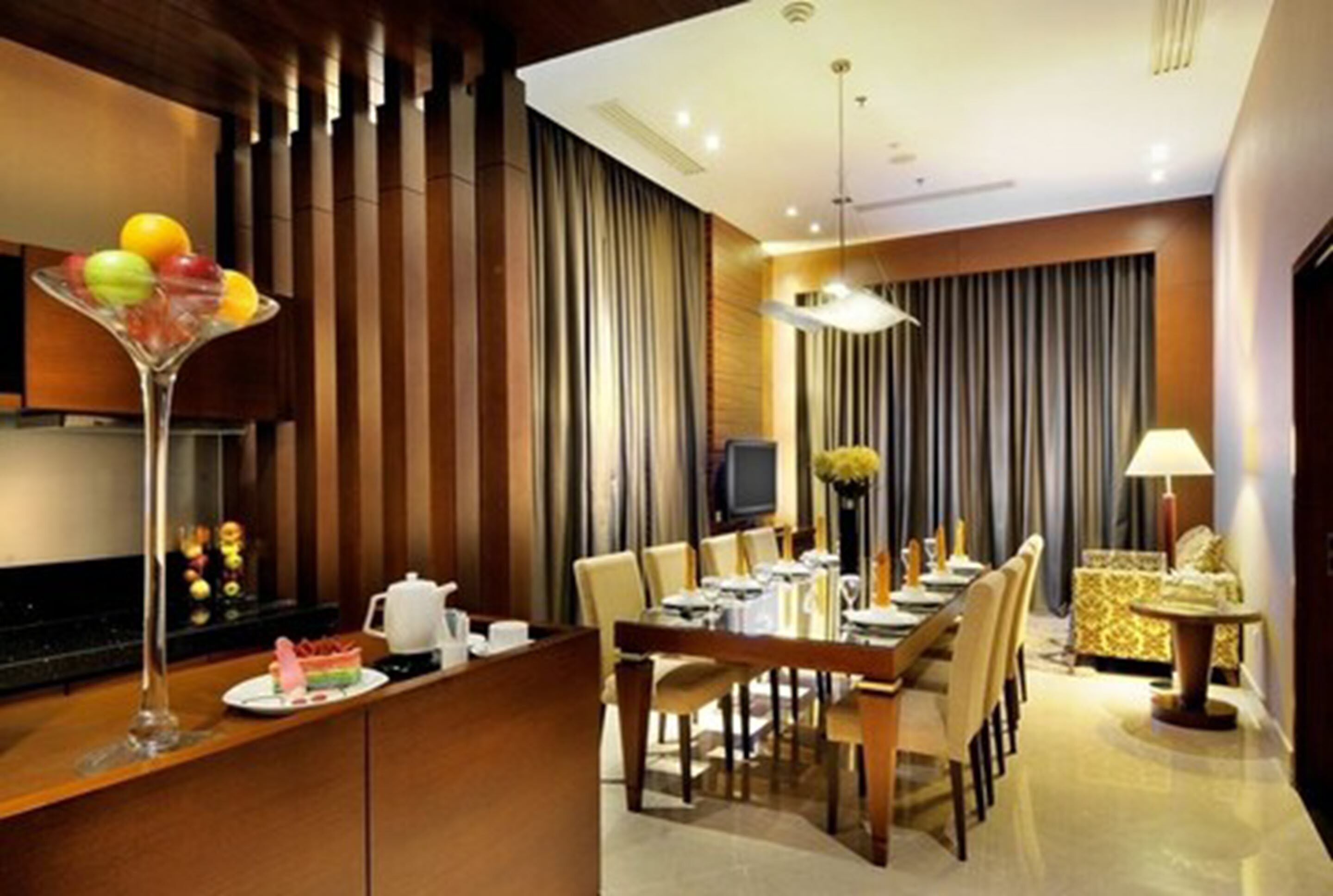 Un comedor moderno de hotel con una larga mesa dispuesta para varios invitados, una zona de bar con una copa de martini, y un televisor al fondo.