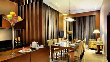 Un comedor moderno de hotel con una larga mesa dispuesta para varios invitados, una zona de bar con una copa de martini, y un televisor al fondo.