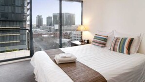 A hotel room with a large bed, a nightstand, a lamp, and a view of the city through large windows. - Inner Melbourne Serviced Apartments (Southbank)