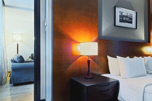 A hotel room with a bed, a nightstand with a lamp, a framed picture on the wall, and a view of another room with a sofa and lamp.