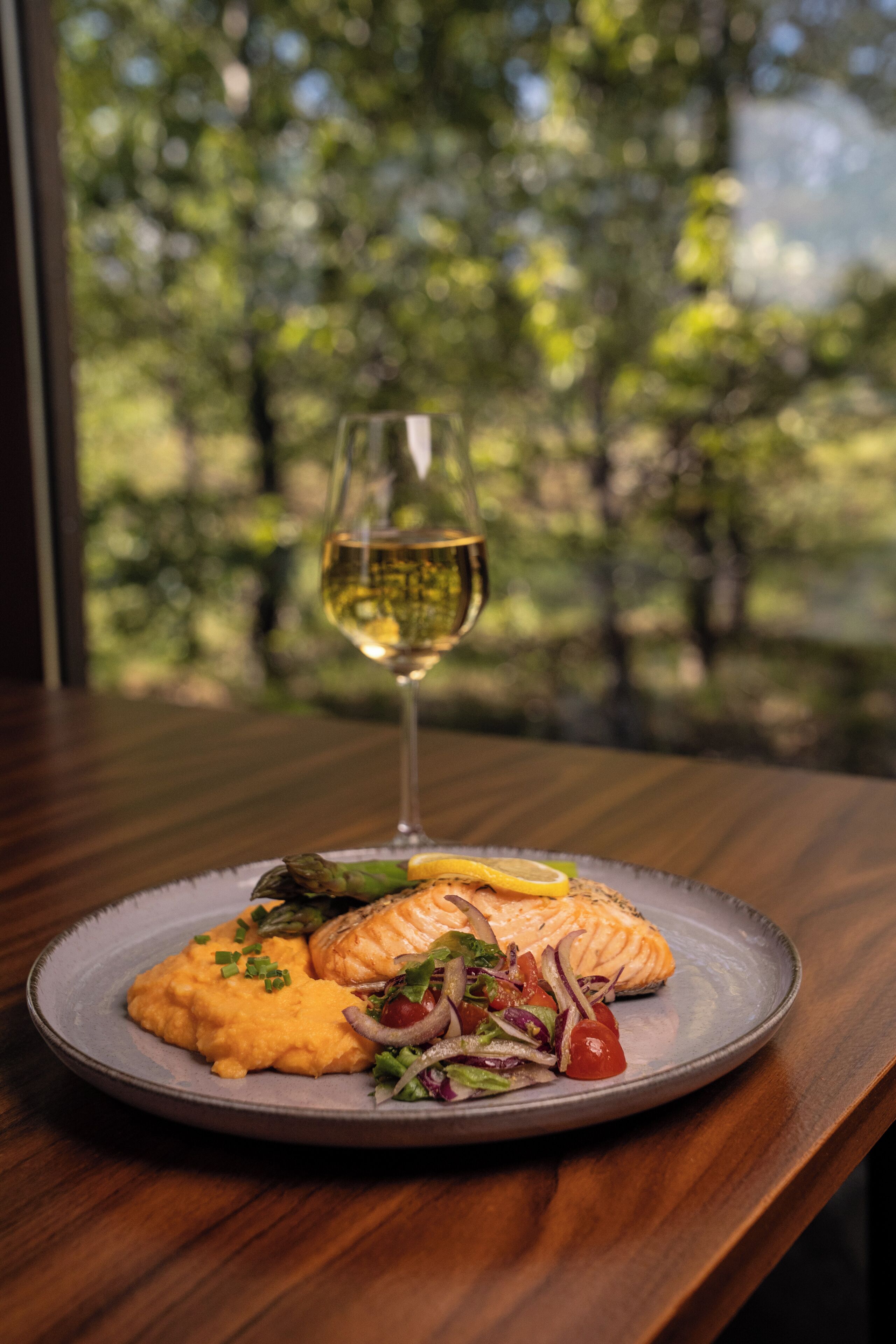A plate of food with a glass of white wine on a wooden table with a view of trees.