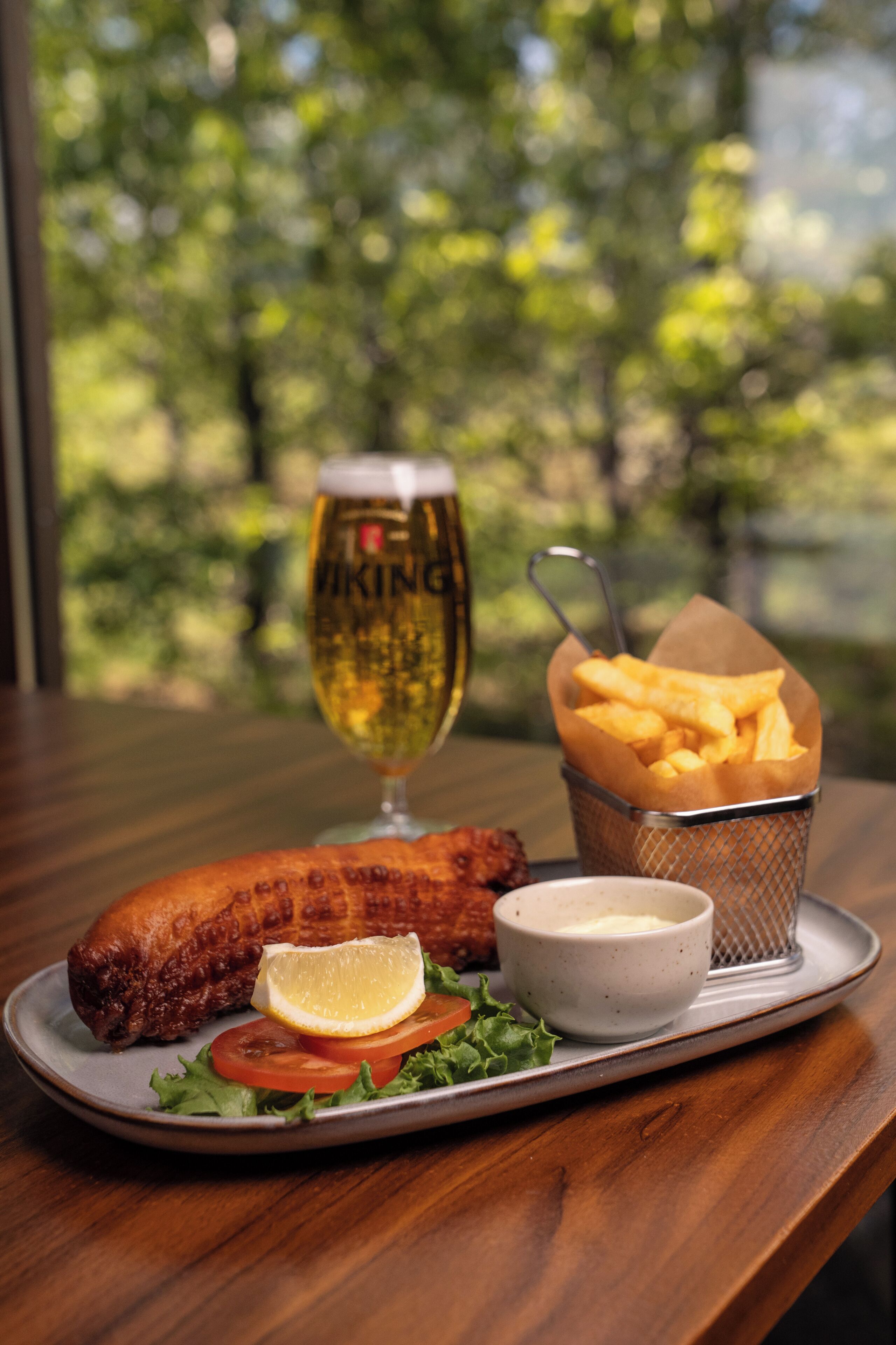 Un plato con pescado, papas fritas, una jarra de cerveza y un recipiente pequeño con salsa, sobre una mesa con vista a los árboles.