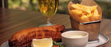 A plate with a fish, fries, a glass of beer, and a small bowl of sauce on a table with a view of trees.
