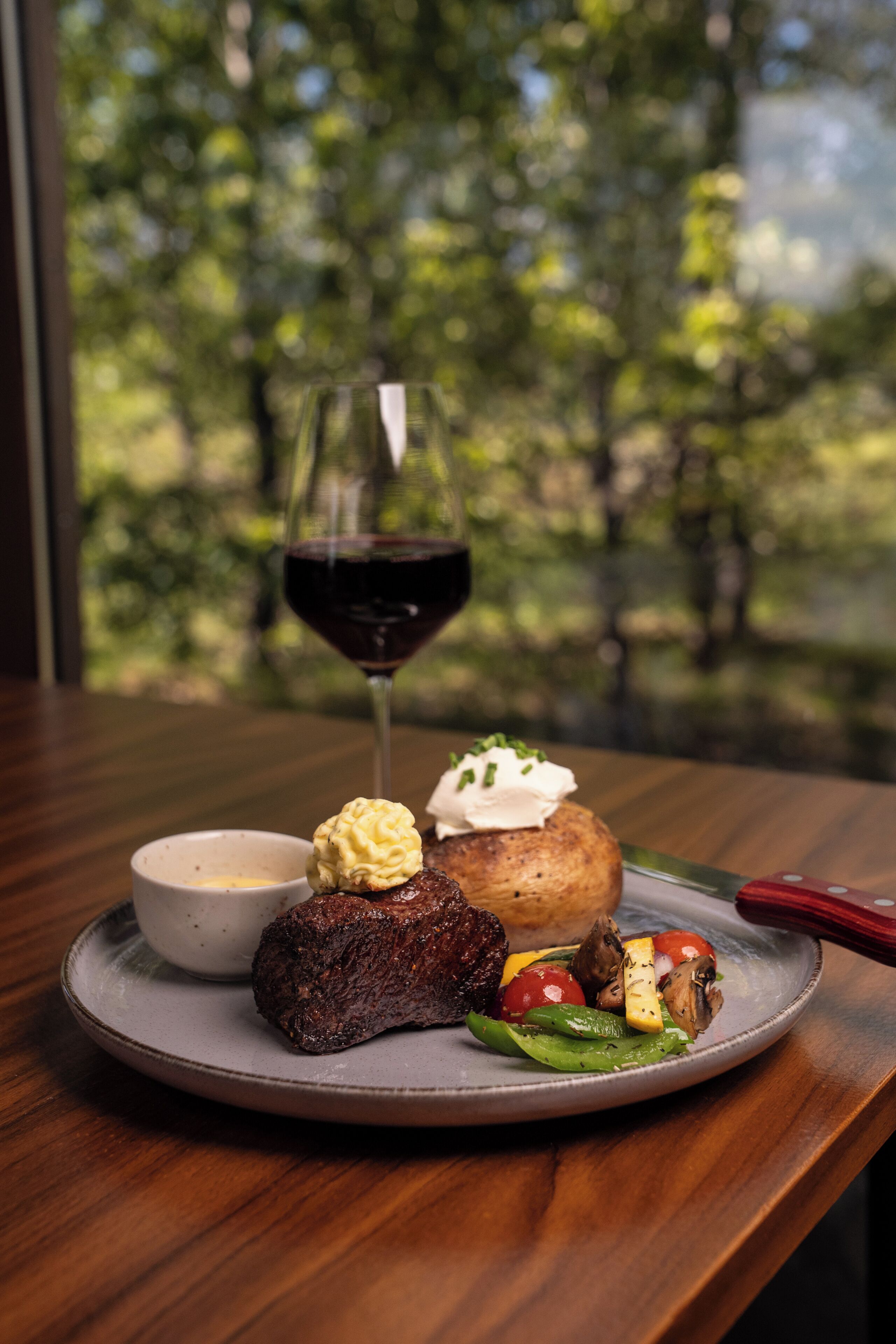 Une assiette remplie de nourriture et un verre de vin rouge sont posés sur une table, avec une vue sur les arbres.