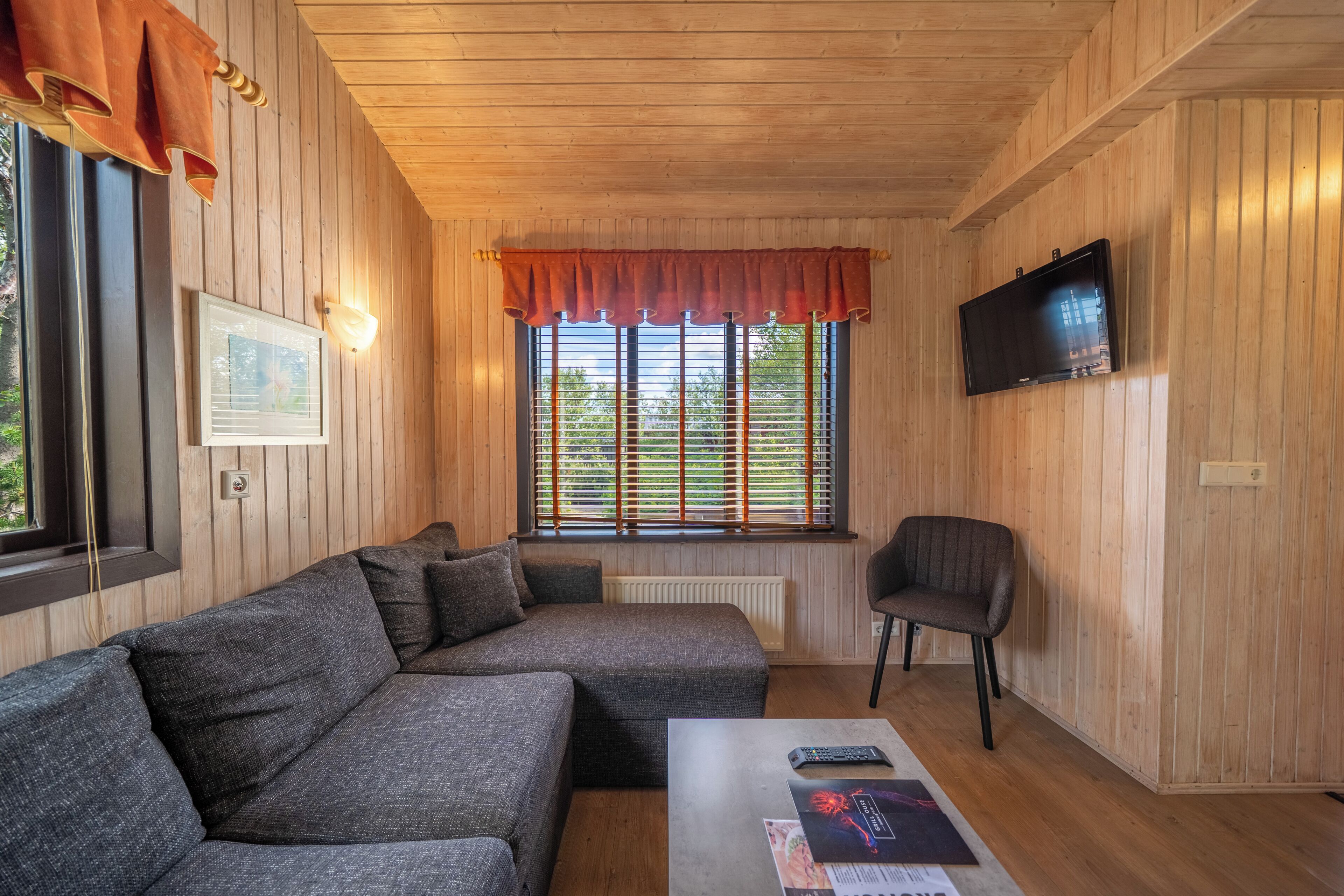 Una habitación con paneles de madera, un sofá modular, una butaca, un televisor de pantalla plana y una ventana con persianas.