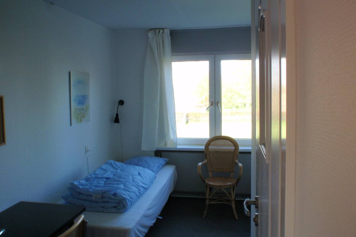 A small, sparsely furnished room with a single bed, a chair, a desk, and a window with curtains.