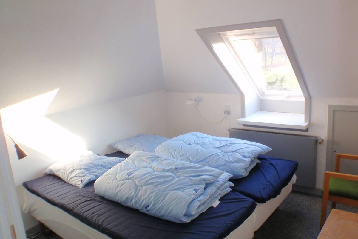 A single bed with a skylight, a bedside table, and a chair in a room with white walls.