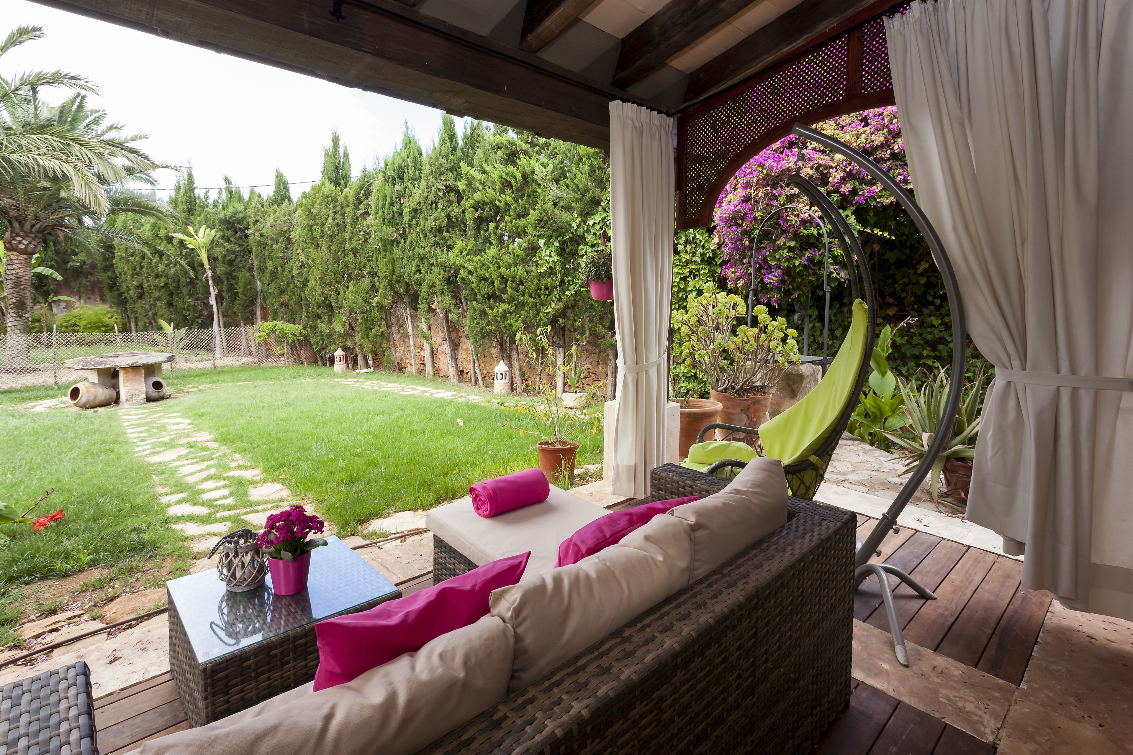 Un espacio de estar al aire libre cubierto con mobiliario de mimbre, una mesa con un jarrón de flores y vistas a un jardín exuberante con un sendero que lleva a un pabellón.
