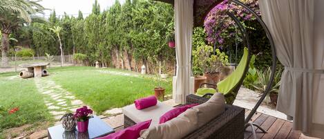 Un espacio de estar al aire libre cubierto con mobiliario de mimbre, una mesa con un jarrón de flores y vistas a un jardín exuberante con un sendero que lleva a un pabellón.