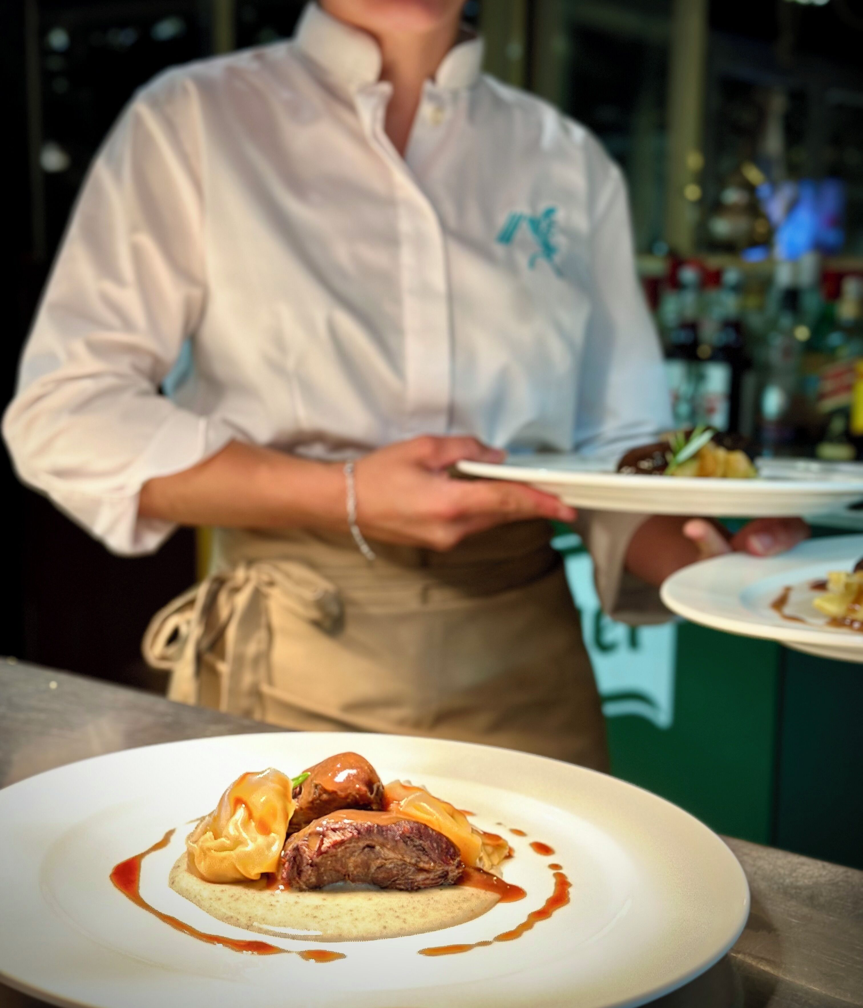 Un xef aguantant un plat amb un plat que conté carn i salsa, amb un fons borrat d'una zona de bar.