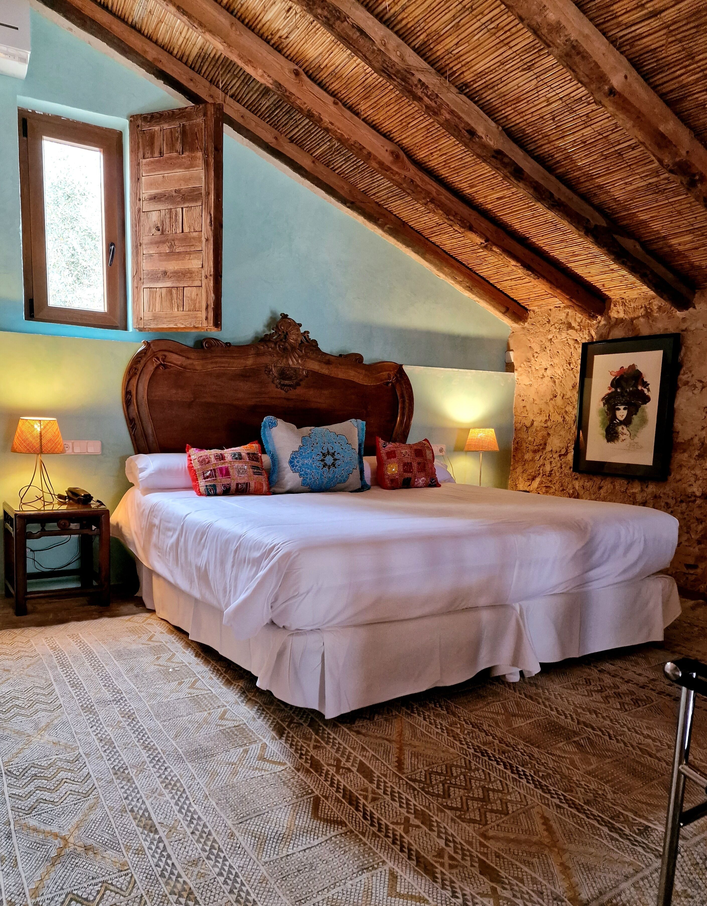 A bedroom with a wooden ceiling, a bed with white bedding and colorful pillows, a nightstand with a lamp, and a framed picture on the wall.