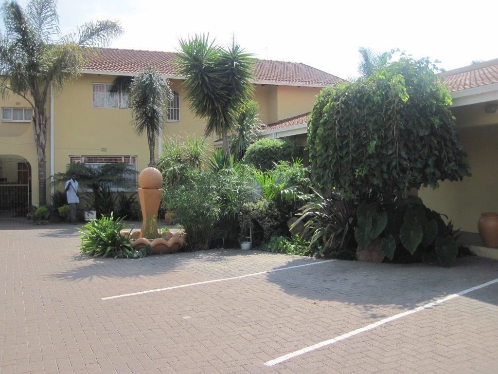 A courtyard with a paved walkway, a yellow building with a red-tiled roof, and various tropical plants.