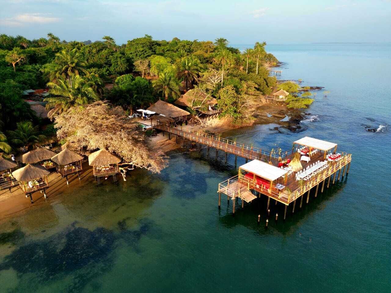 Un muelle se extiende sobre un mar tropical con construcciones de techo de paja y un área de comedor.