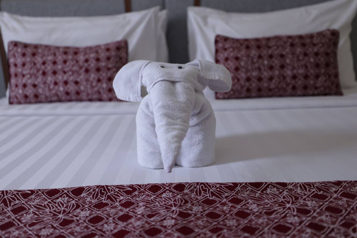 Una cama bien hecha con una colcha blanca y roja estampada, dos almohadas blancas y una toalla blanca con forma de elefante.