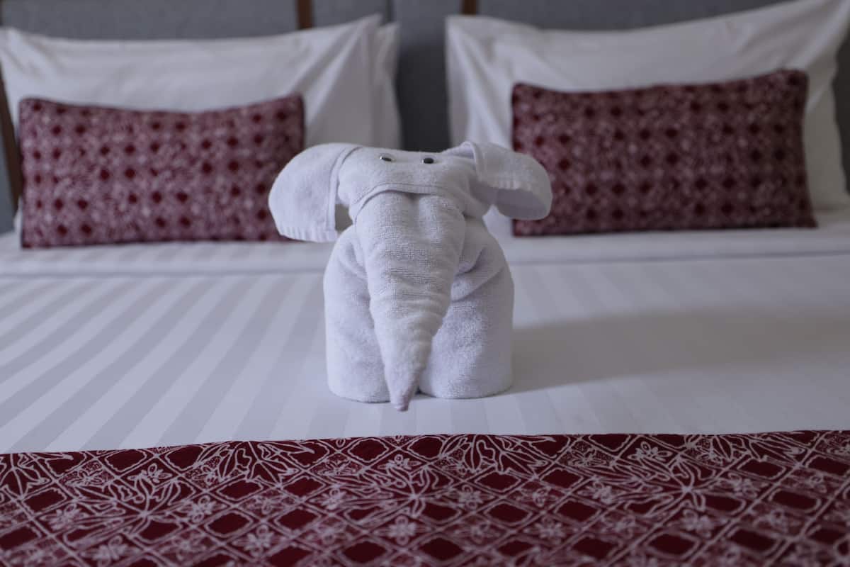 Un lit bien fait, recouvert d’une courtepointe blanche et rouge à motifs, avec deux oreillers blancs et une serviette blanche en forme d’éléphant.