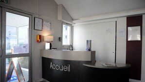 A reception desk labeled "Accueil" with a computer, a telephone, and a small table. There are glass doors leading outside, and a fire extinguisher is mounted on the wall. - Hôtel Inn Design & Restaurant Amiens (Dury)