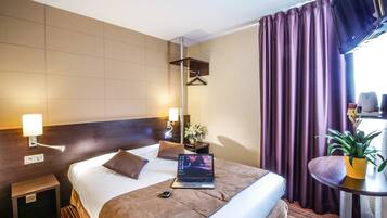 A hotel room with a bed, bedside table, lamp, laptop, and a small table with a plant.