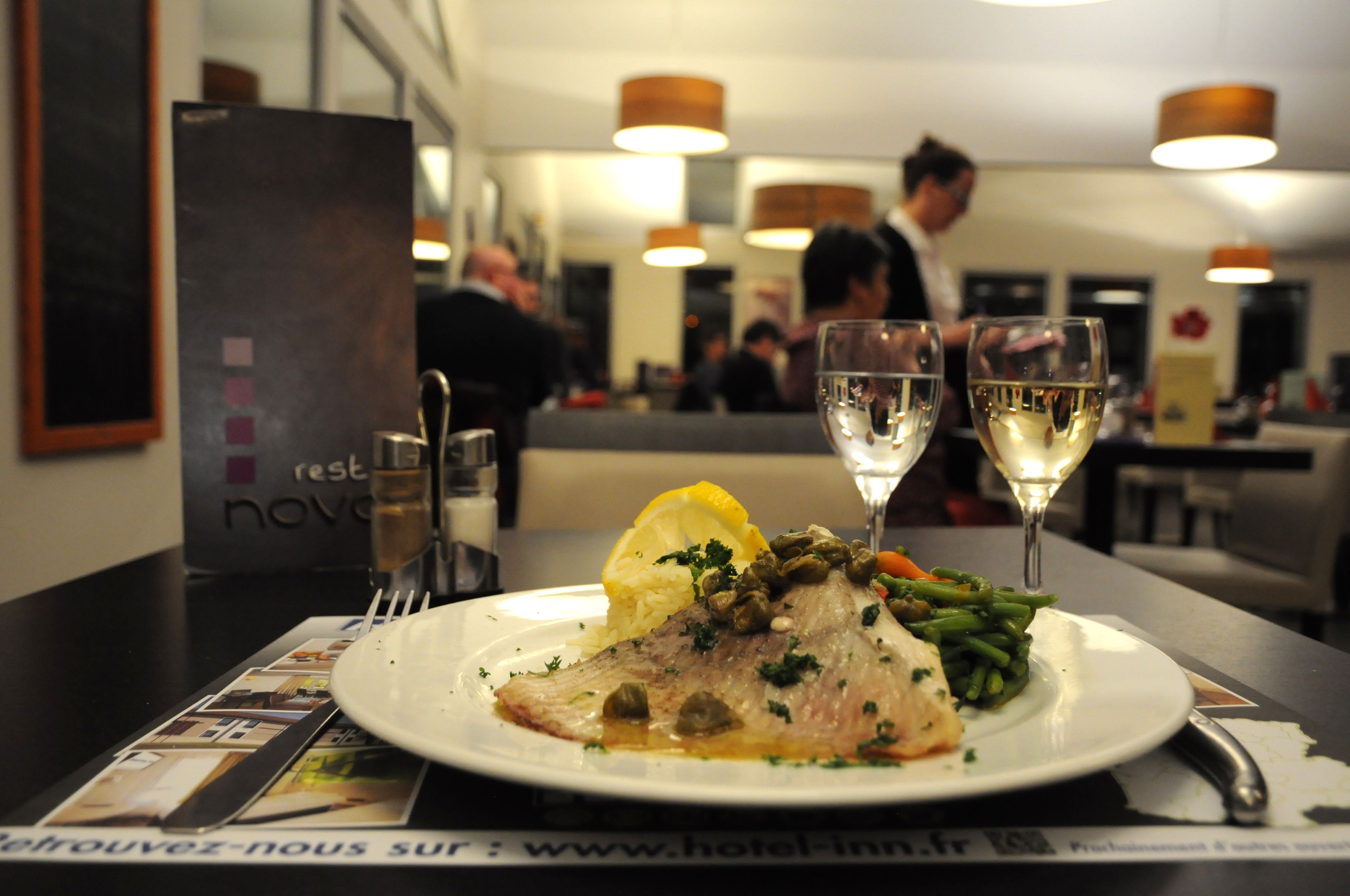 Une table à manger avec une assiette de nourriture, deux verres de vin blanc et un menu.