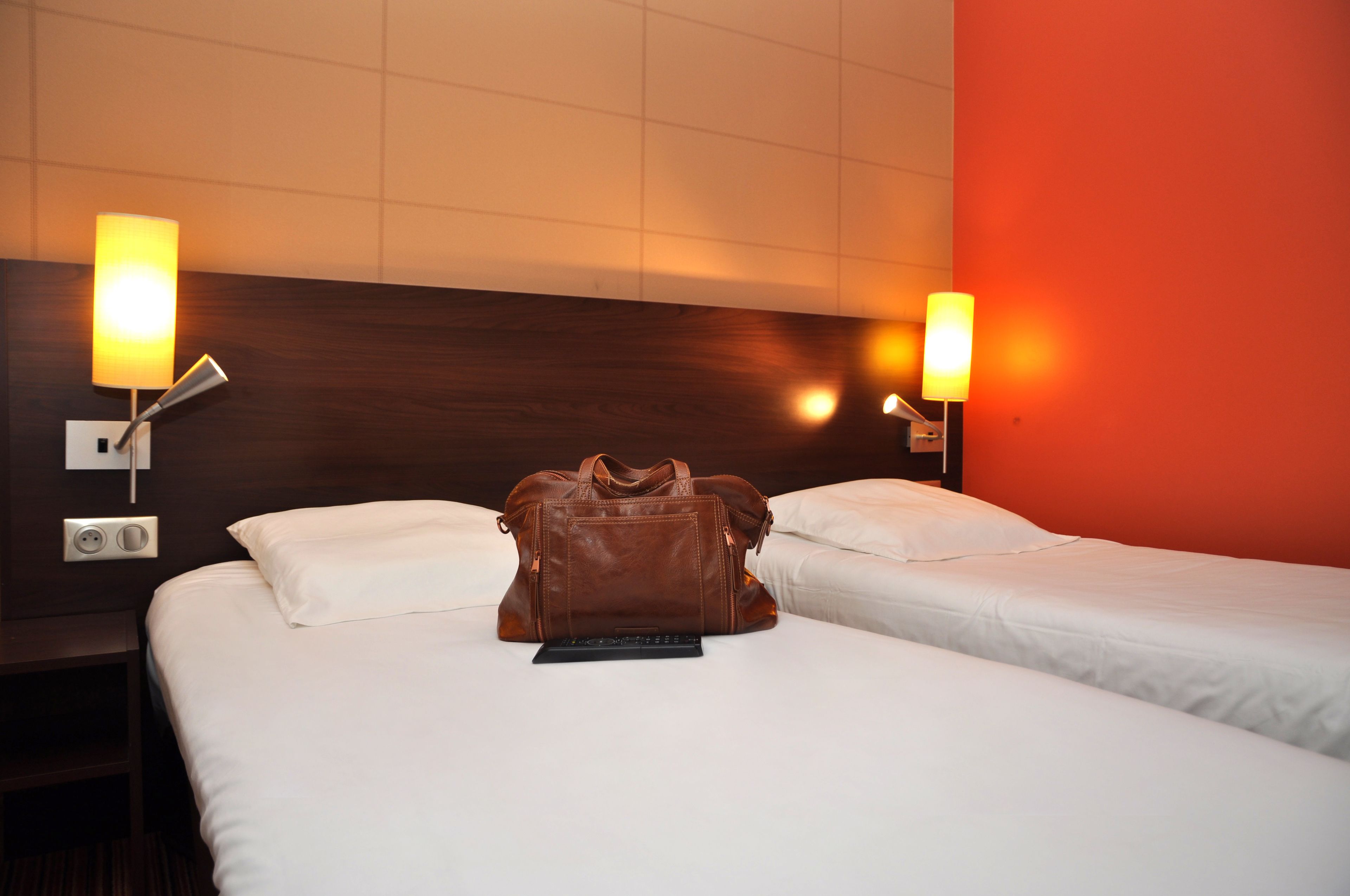 A hotel room with two beds, a brown leather bag on the bed, and wall-mounted lamps.