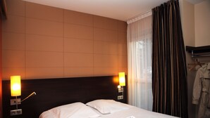 A hotel room with a bed, two bedside lamps, a robe hanging on a rack, and a window with curtains. - Hôtel Inn Design & Restaurant Amiens (Dury)