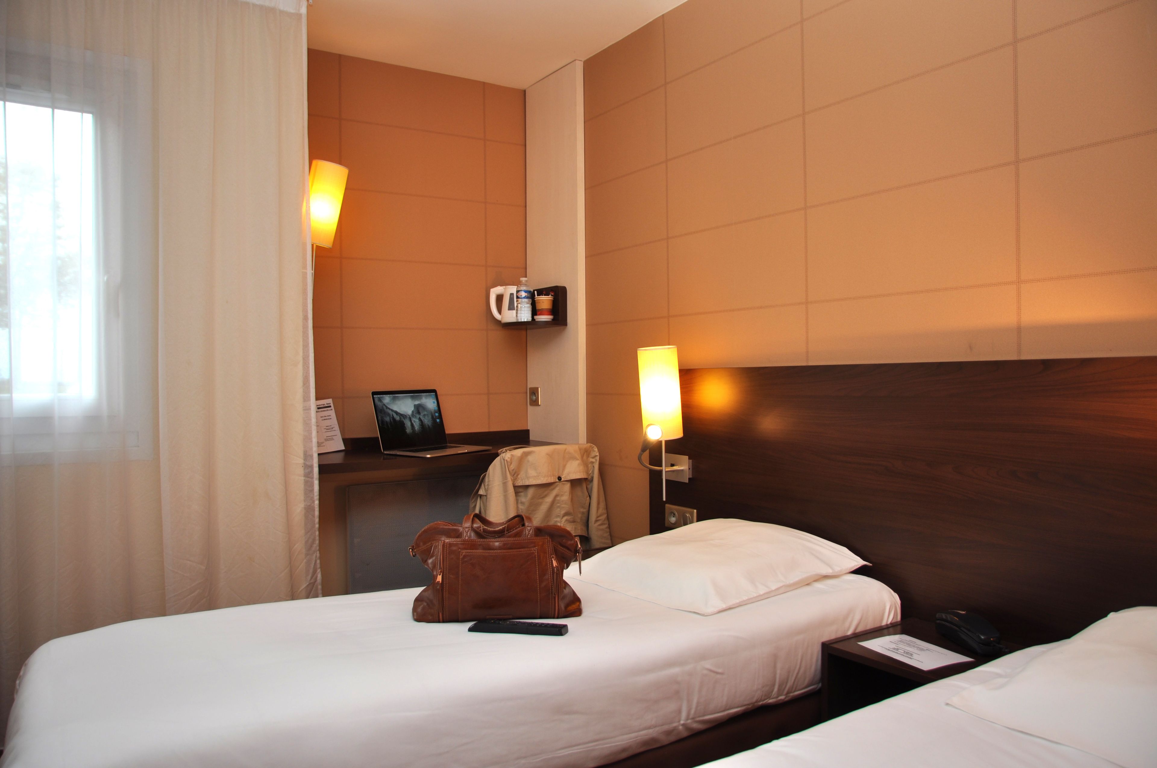 A hotel room with two beds, a desk with a laptop, a coat rack, and a wall-mounted shelf with a coffee maker and a mug.